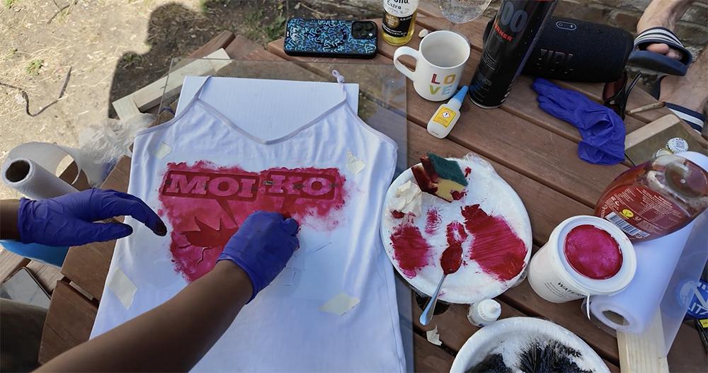 A person wearing gloves painting the 'Moi Ko' logo on a tank top, with objects scattered around the table.