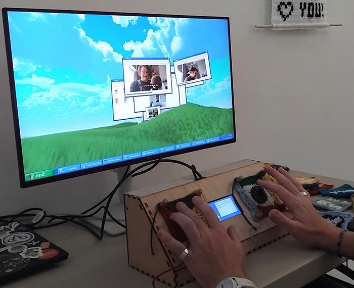 A person's hands using an assistive communication device connected to a monitor displaying a video call.