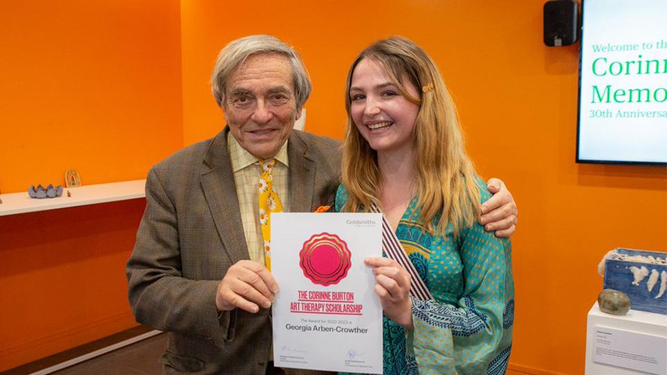 Sir Michael and Georgia Arben-Crowther stand facing the camera holding a certificate between them