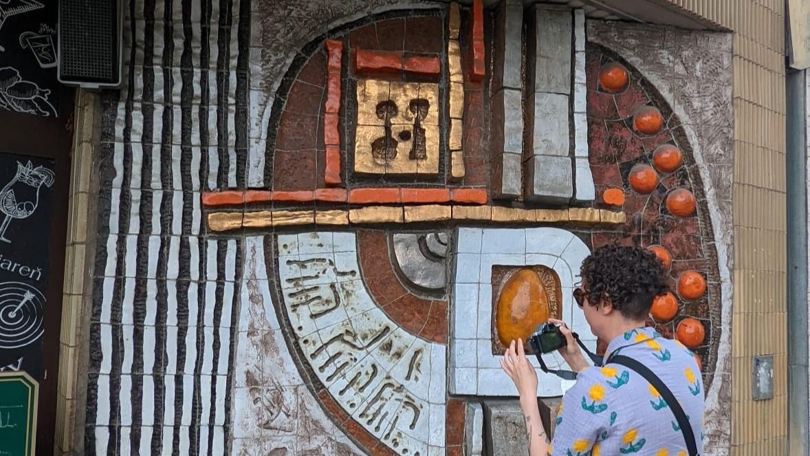 Vik Loveday stands in front of Kosice mosaic to take a picture with a digital camera