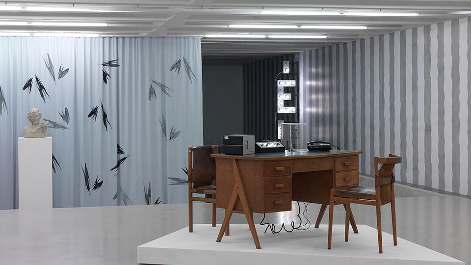 Installation with mid-century desk and chair on a raised white platform in front of a striped wall and light grey curtain.