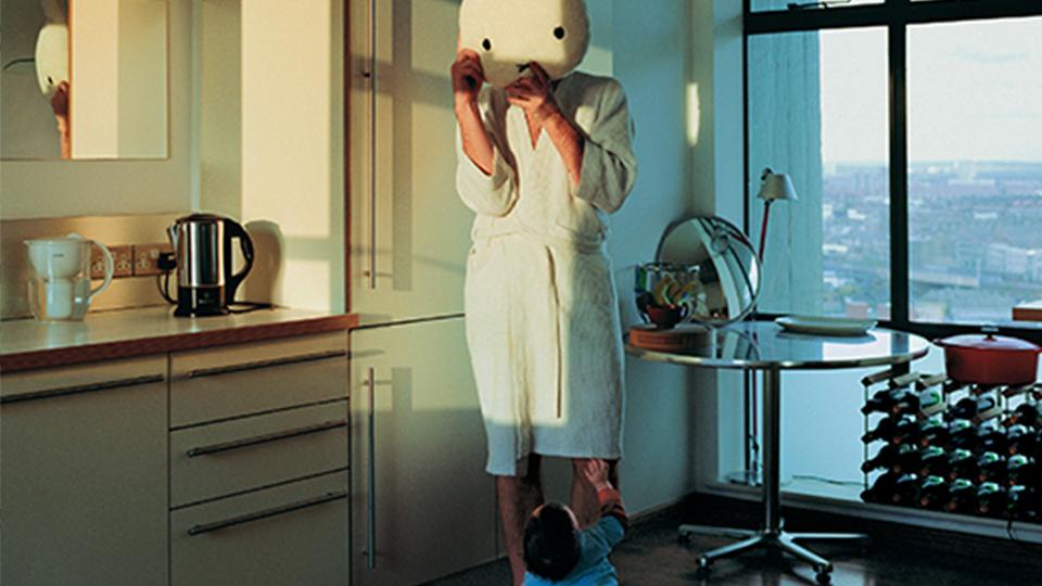 Photograph of person in dressing gown wearing a Hello Kitty head, standing in a kitchen with a toddler at their feet.