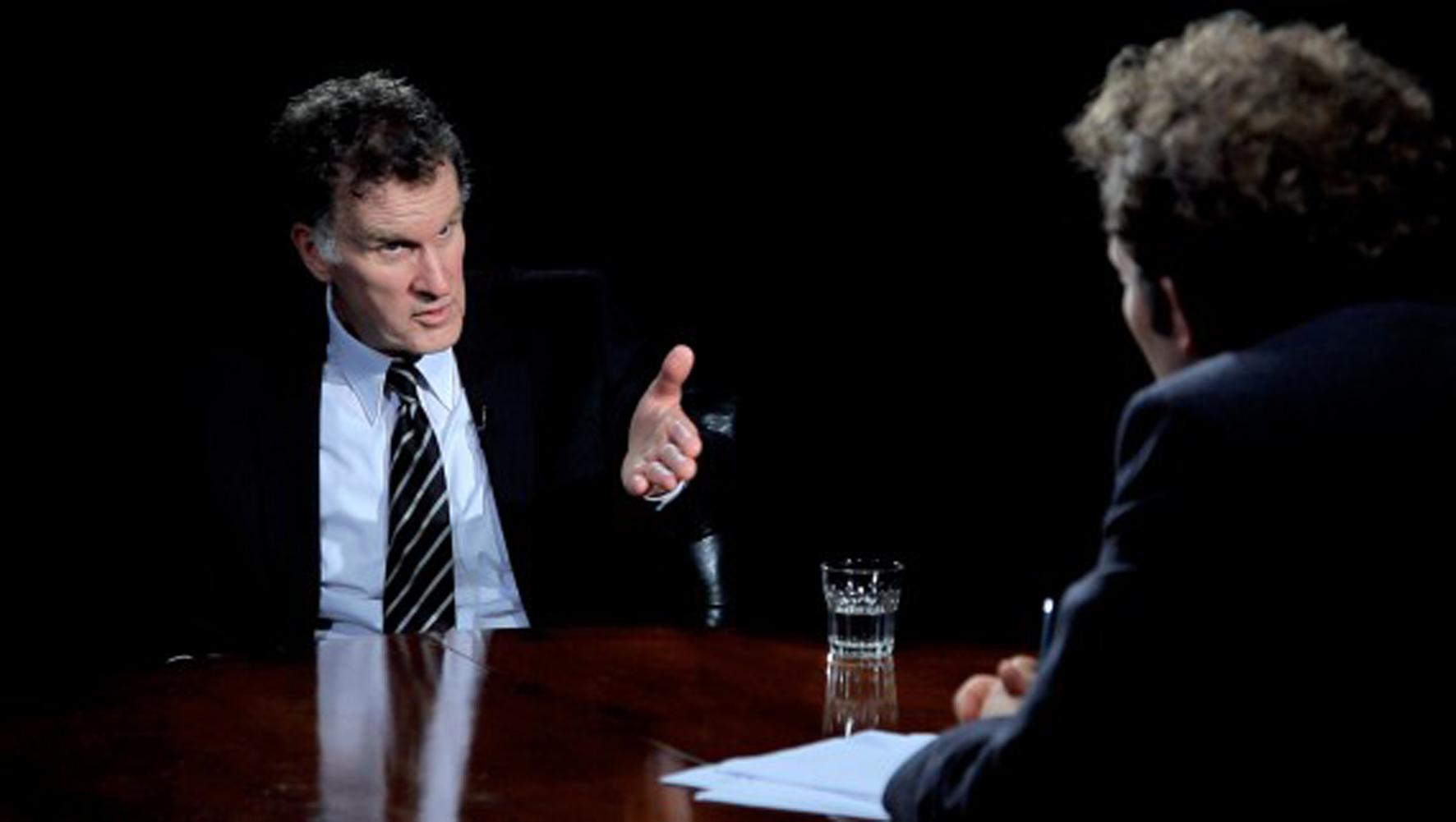 Two actors sitting at a table staging an interview between a banker and a host. A video still from a work by Jan Peter Hammer