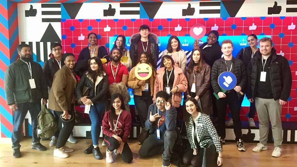 A large group of students stand in front of a colourful wall at Facebook's office.