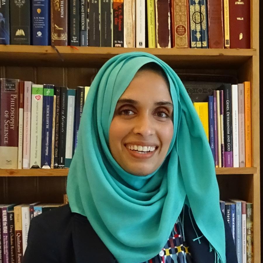 A woman wearing a bright blue headscarf stands smiling in front of a bookcase