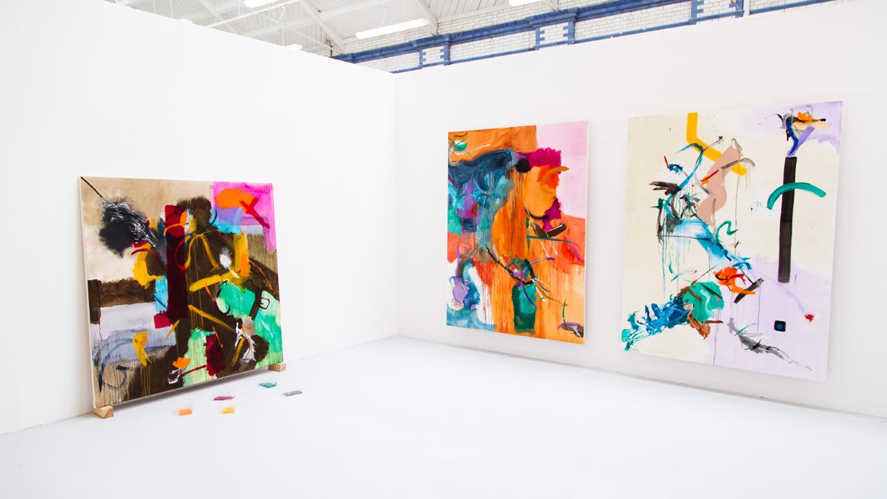 Three large, colourful, abstract paintings hang on white walls in the corner of a gallery.