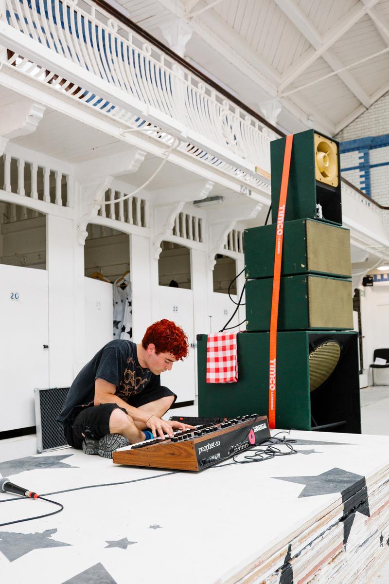 Performer with red hair playing a synthesiser on a painted stage beside stacked green speakers in a white hall