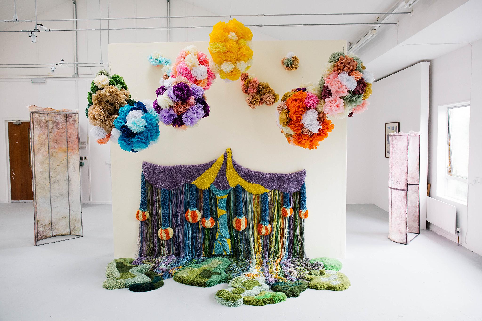 Colourful textile art in a gallery featuring a circus tent design with hanging yarn, pom-poms, moss-like rugs, and floating fabric flower clusters above