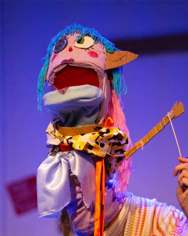 A scrappy looking puppet with blue hair and mismatching eyes, its mouth is open
