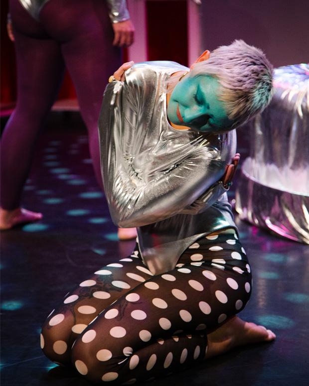 A woman in a silver costume is crouched on the floor with her arms across her body, her face is painted green and she has silver hair