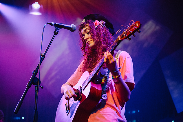 A woman in a pink top and black beret playing guitar and singing into a microphone