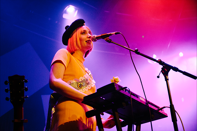 A woman in a yellow dress and beret wearing a sash and playing the keyboard and singing into a microphone