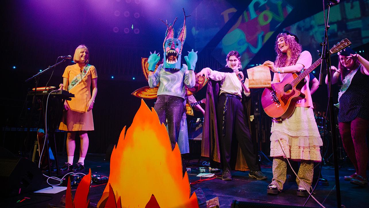 The band on stage around a cardboard campfire. There is a person dressed up as a blue alien, and another dressed up as a vampire, and two women dressed as girl scouts - one has a guitar, and one is stood by a keyboard. There are other bandmates in the background. 