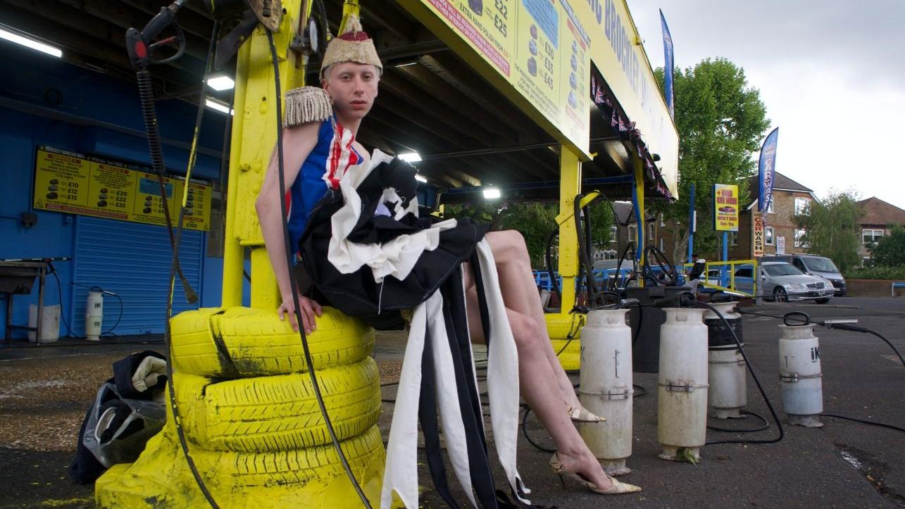 A man sitting on a stack of tyres painted yellow outside a car wash wearing epaulettes and a military style hat, black and white skirt and heels. 