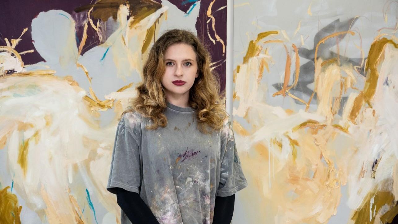Michaela, wearing a grey T shirt with paint stains, stands in front of one of her paintings. 