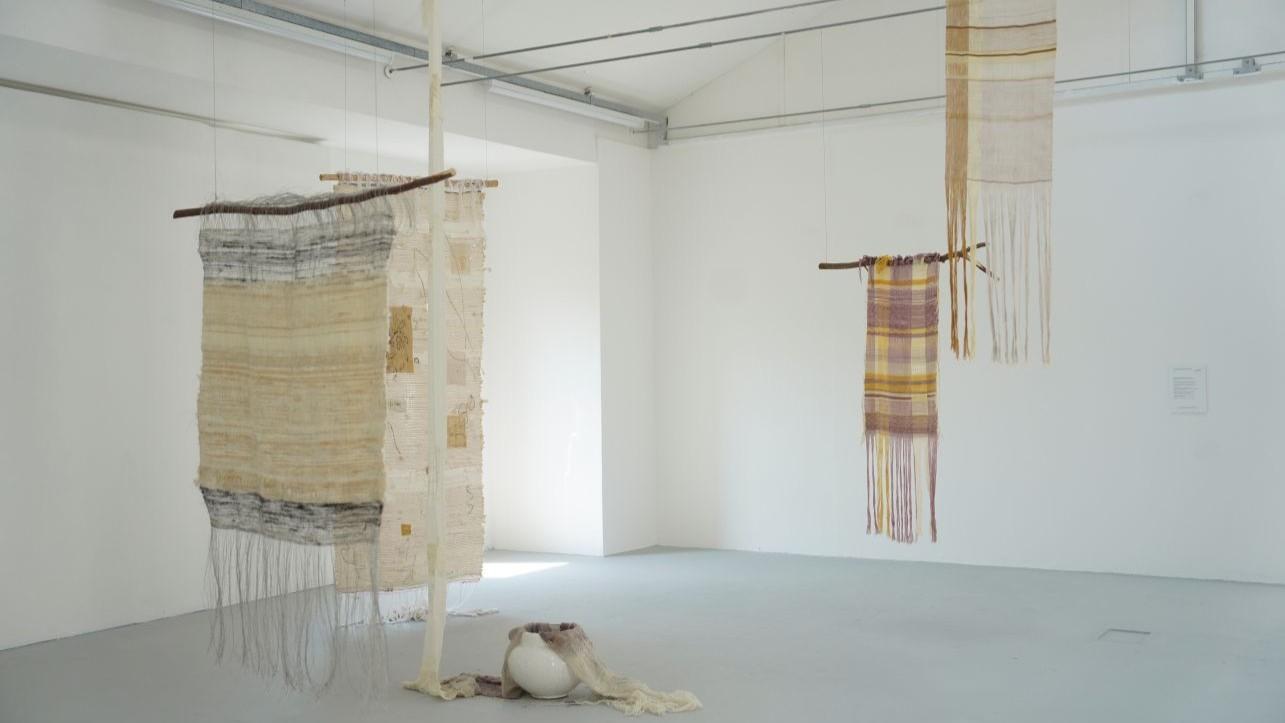 Five weavings hanging from the ceiling in a space with white walls. One is dipped into a white pot of blueberry dye. 