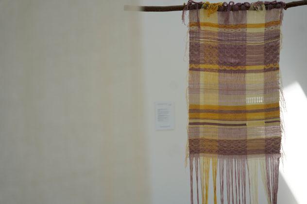 A weaving with purple and yellow stripes suspended from a branch, with another off-white weaving close up in the foreground