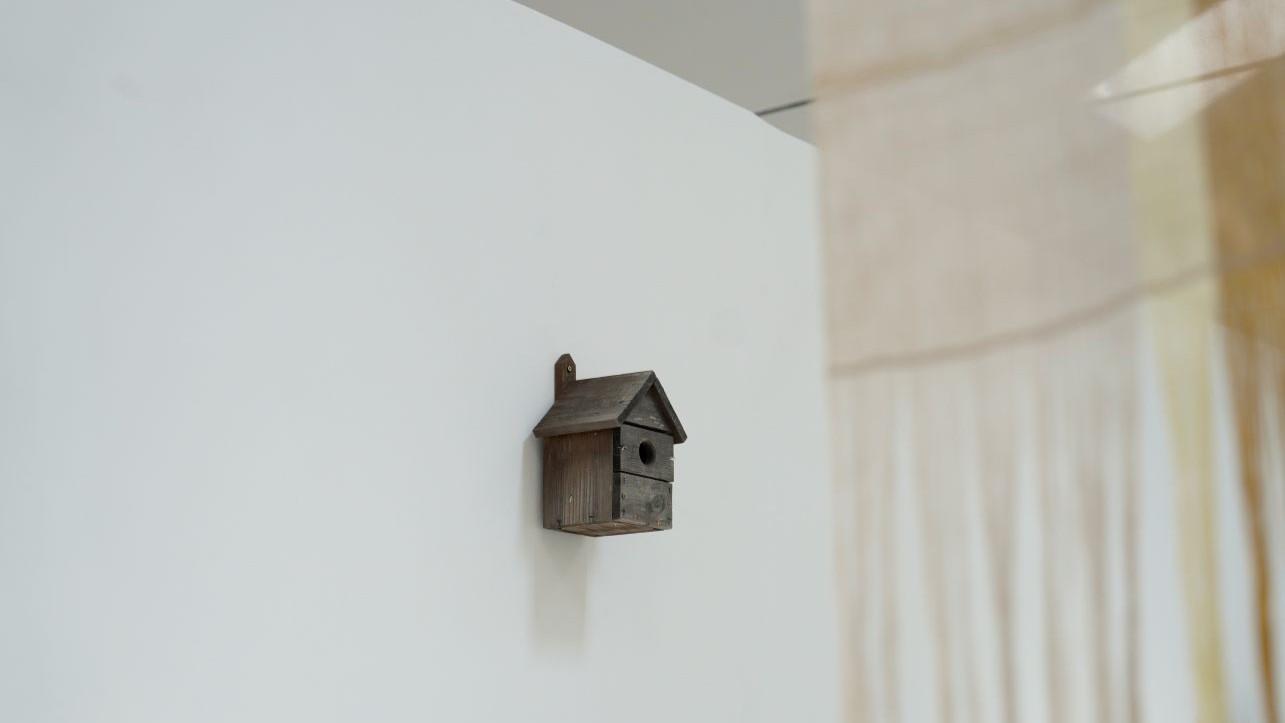 A wooden bird box installed on a white wall, with a weaving out of focus in the foreground