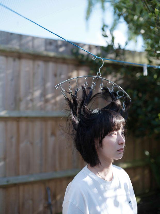A photograph of a woman with a coat hanger with pegs on it attached to her hair, attached to the washing line, with a wooden fence behind. 