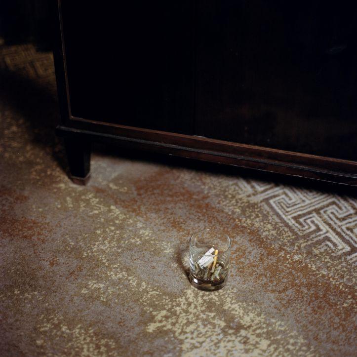 A photograph of a glass on a carpeted floor, with cigarette butts in it.