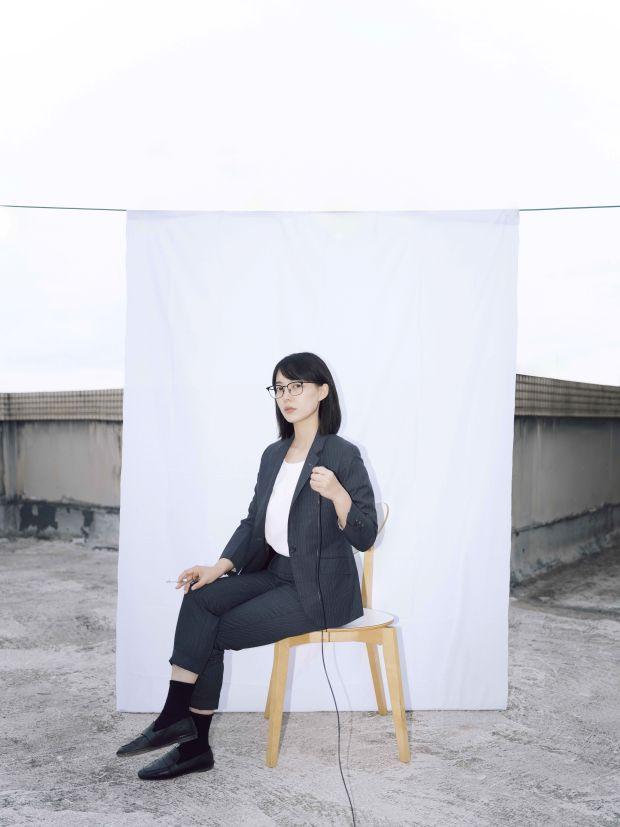 A woman in a suit wearing glasses sits on a wooden chair in front of a white sheet.