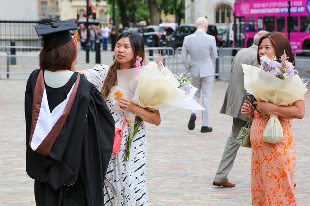 Supporters stand with bunches of flowers, one helps fix a student in a graduation robe's hair