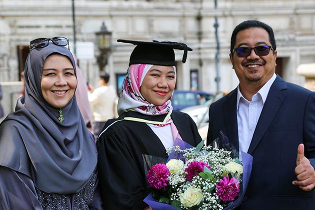 A female graduate stands between her two parents. Her father has his thumb sticking up.