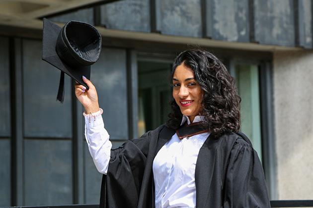 A female graduate stands outside holding her hat up to her right in salute.