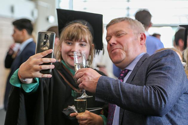 A female graduate and her father pose for a camera phone photo holding up their glasses. The father has a proud smile on his face.