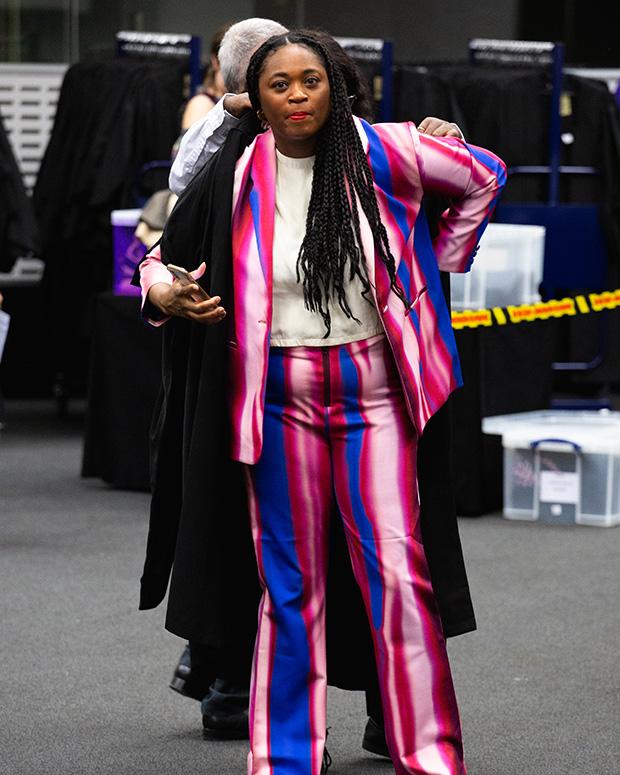 A graduand in a bright purple and pink stripped suit gets her gown fitted on top of it