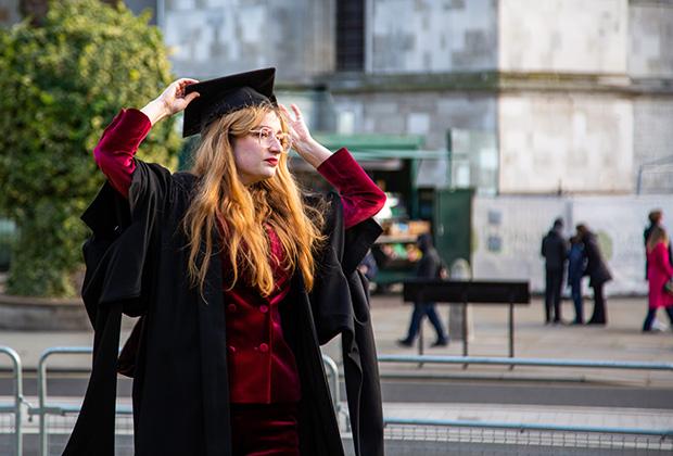 A graduate in a dark red jacket adjusts her mortar board outside the venue.