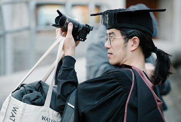 A graduate outside holding up a camera to take a photo while carrying a large baf