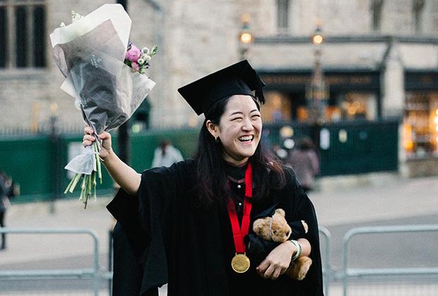 A graduate outside holding a bunch of flowers aloft with her right hand. In her left hand is a teddy bear.