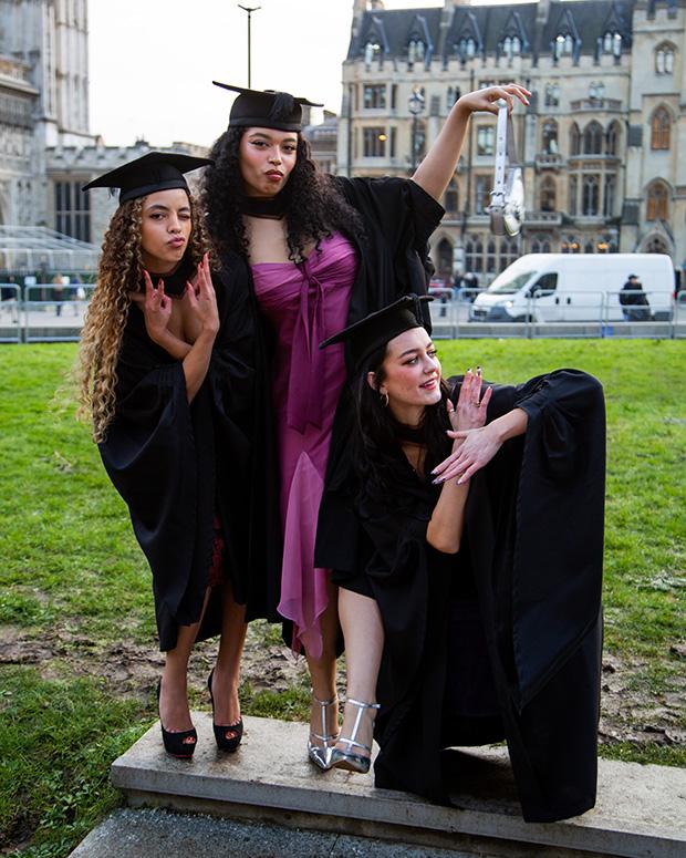 Three graduates pose outside each making shapes with the arms and pointing
