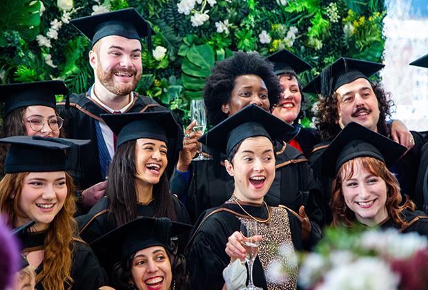 10 graduates pose as a group against a green wall all smiling