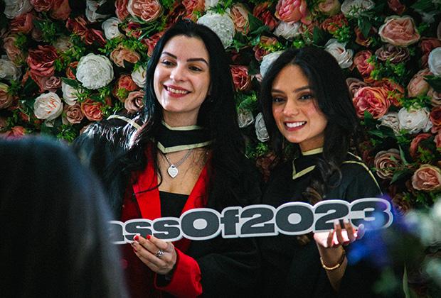 Two graduates next to a flower wall holding a 'Class of 2023' sign