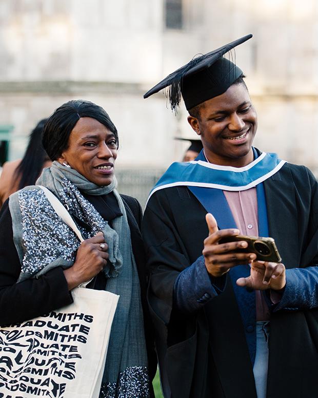 A graduate looks at his phone next to his guest who is carrying a graduation tote bag