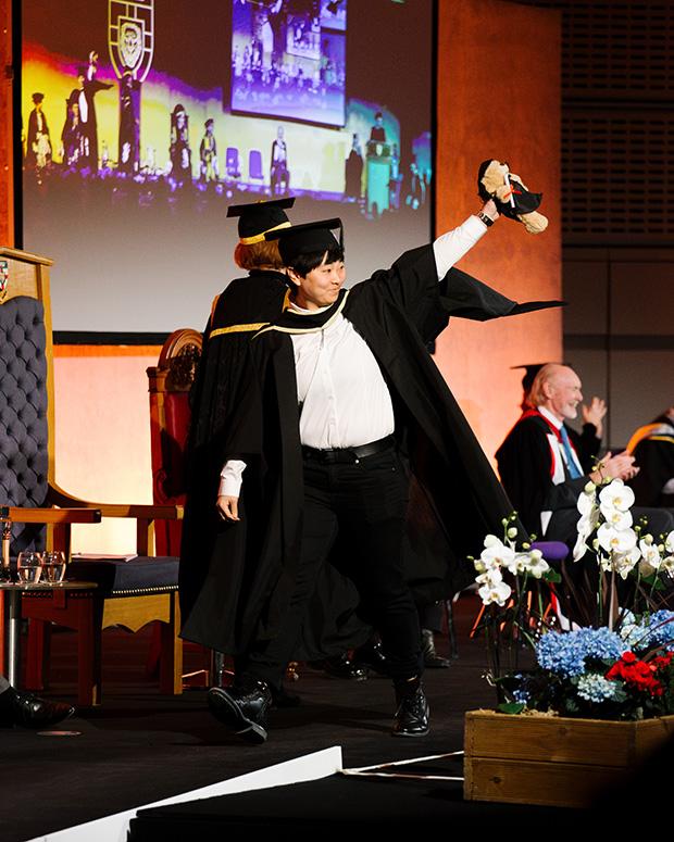 Graduate holds a teddy in the air as they walk across the stage