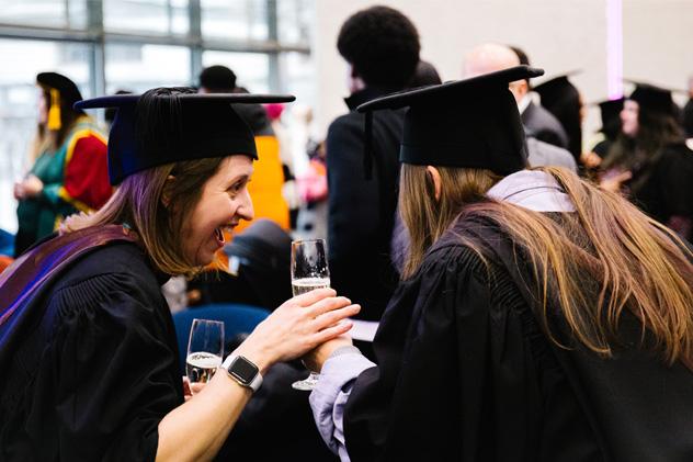 Two women in graduation gowns laugh at a reception.