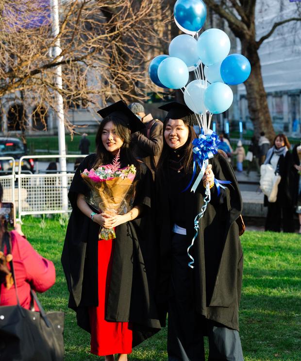 Two graduates pose for photographs outside with blue balloons behind them 