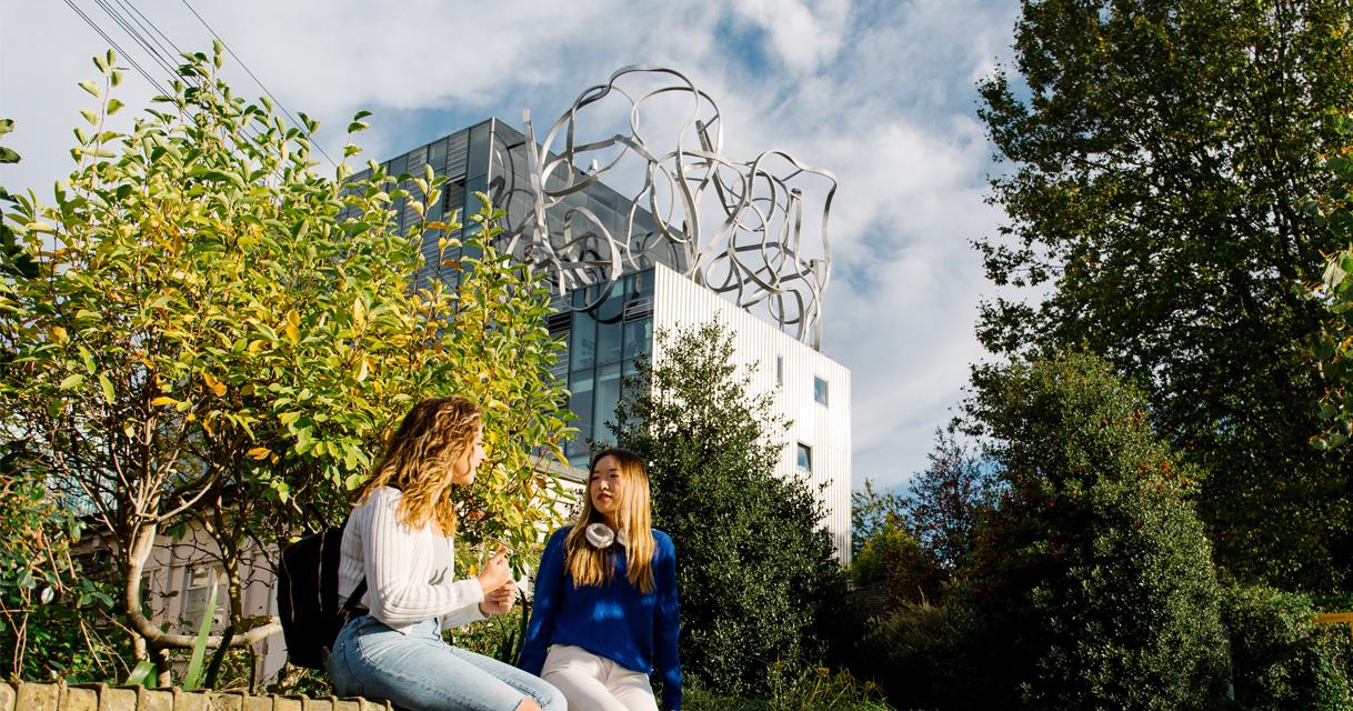 Two students sit chatting with Goldsmiths' Ben Pimlott Building in the background.