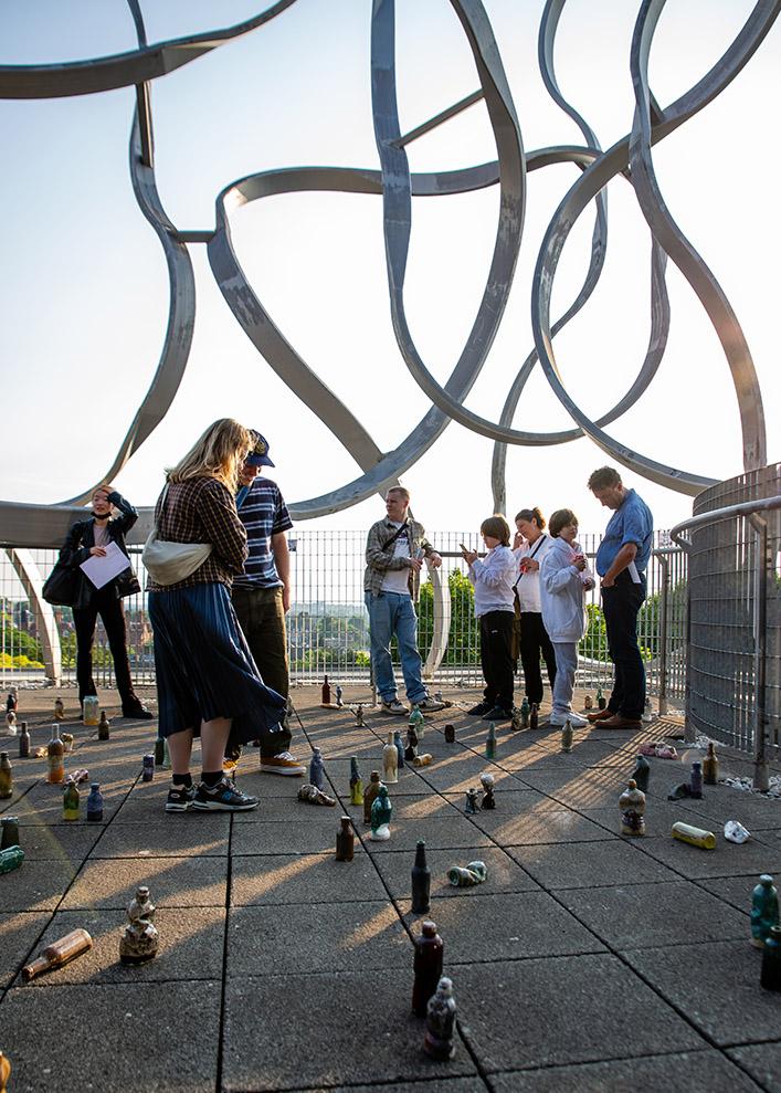 A group looking at artwork on the ground. Above them is the Squiggle, a metal sculpture made of a continous string of metal.