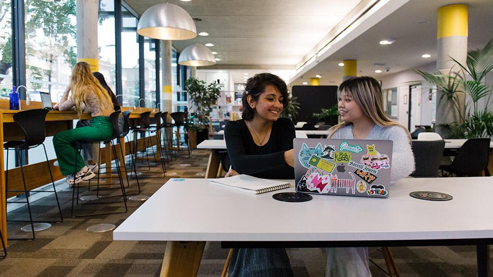 Two students working together on a laptop at a desk in the Library