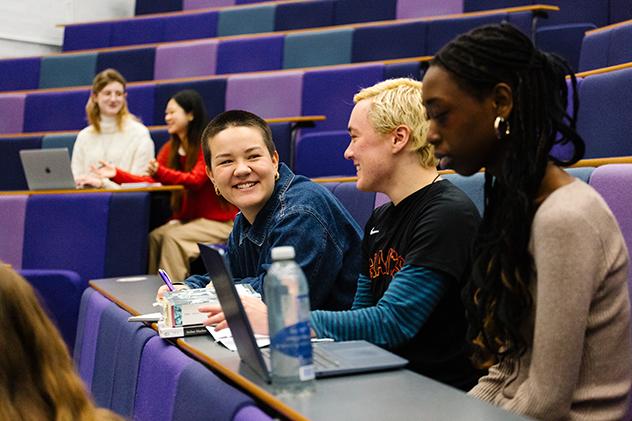 Students sit in a tiered lecture hall with purple seats, working on laptops and talking in small groups. In the foreground, three students sit together at a desk; one smiles toward a classmate while notebooks, laptops, and a water bottle are visible.