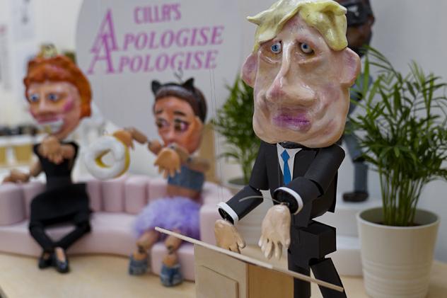 BA Design student work from the 2022 degree show, depicting a Boris Johnson puppet in front of a plinth, with a sign in the background saying 'Cilla's Apologise Apologise', and two other puppets sitting on a pale pink sofa in front of it
