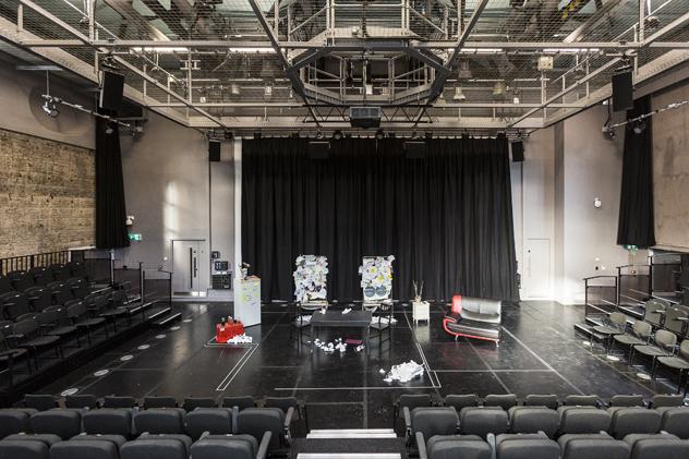 A wide view of the on-campus George Wood Theatre, with seating to the foreground and each side, props including a sofa and table on the stage foor, and a lighting rig