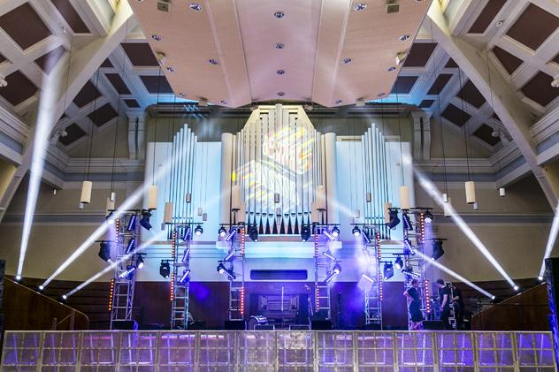 The Goldsmiths Great Hall, which has vaulted ceilings and a stage that features a grand organ in the background. The stage features music festival lighting.