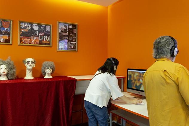 An orange room displays work from final-year media and communications undergraduates, including three papier mache heads, and iMacs that visitors listen to with headphones