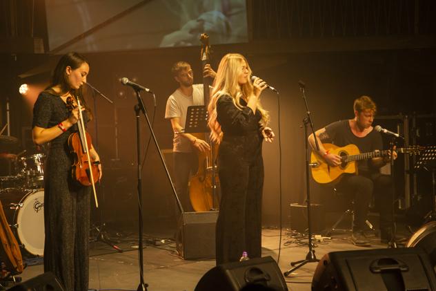 A four-person band performs at Goldsmiths' annual music festival, PureGold. The band features someone singing into a mic, with three bandmates holding strings instruments – a violin, cello, and acoustic guitar.
