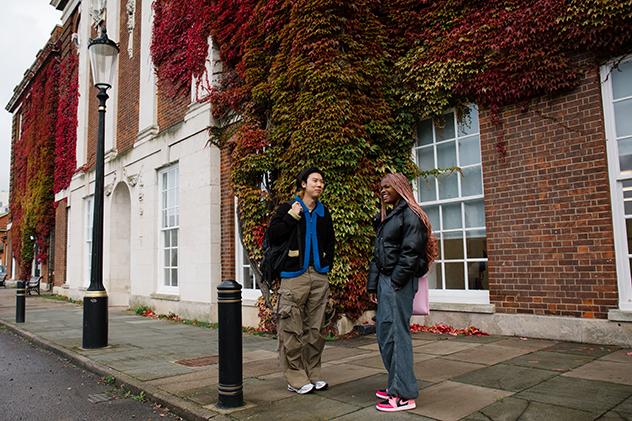 Two students standing and talking beside the Richard Hoggart Building, which is covered in dense red, orange, and green ivy.
